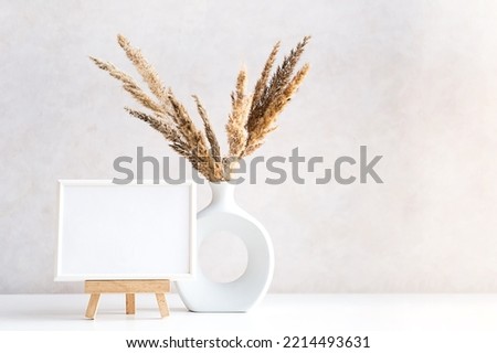 Empty picture frame mock up in minimalist interior with dried plant in vase on white table.