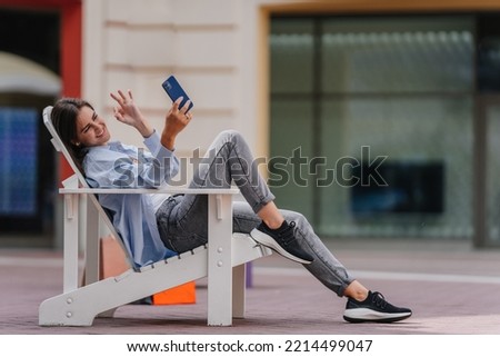 Brunette young woman sitting outside in wooden chair making video call, shows ok sign. Young blogger makes selfie smiling shows ok sign at camera of phone. Trendy cheerful woman in casual having break