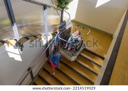 Mother with a young child living with cerebral palsy using electric wheelchair lift to access public building. Special lifting platform for wheelchair users. Disability stairs lift facility. Royalty-Free Stock Photo #2214519029