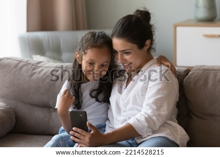 Attractive Indian woman and little cute daughter sit on sofa look at smartphone screen enjoy new mobile apps, calling through video call to family, spend time at home use cellphone. Modern tech, fun