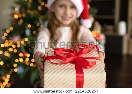 Christmas gift box in little girls hand close up. Sweet preschool child holding, giving, showing at camera festive Xmas present wrap with red ribbon and bow, standing by glowing tree in background