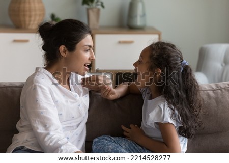 Young Indian mother and cute daughter practicing speaking together, doing vocal exercises seated on sofa at home, therapist logopaedist correct pronunciation of preschooler girl, help to stutter kid Royalty-Free Stock Photo #2214528277