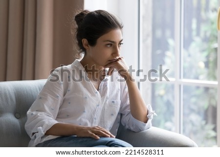Sad Indian woman sit on armchair looks concerned, feels confused, thinks, search solution in difficult life situation, hesitating seated alone at home. Relations problems, break up and marriage split Royalty-Free Stock Photo #2214528311