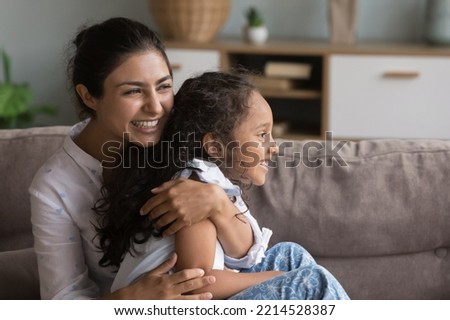 Loving laughing Indian mother hugging her daughter tightly seated on sofa enjoy tender moments spend priceless time together feeling unity and love. Caring parent and children bonding and protection Royalty-Free Stock Photo #2214528387