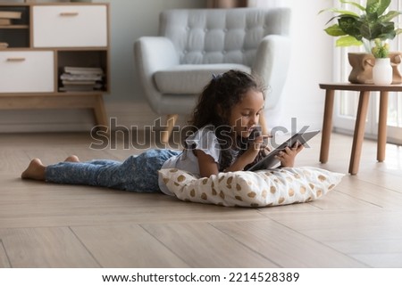 Adorable preschooler little Indian girl lying on warm floor using digital tablet in cozy living room, play videogames, watch favourite vlog, spend time at home. Gen Z use modern wireless technology