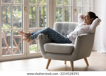 Pretty Indian woman chilling resting on comfortable armchair near panoramic windows in light sunny modern living room, female closing eyes enjoy free time and fresh conditioned air inside cozy house
