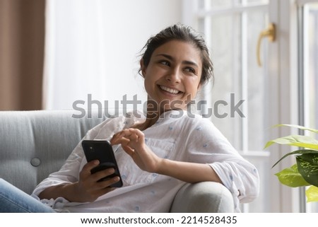 Peaceful Indian woman relaxing on cozy chair with smartphone, smile staring aside enjoy carefree weekend and modern wireless tech. Making call, ordering goods and services on-line, e-date apps usage Royalty-Free Stock Photo #2214528435