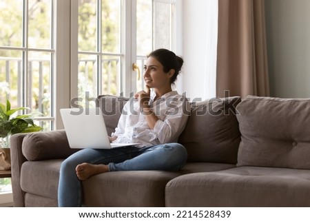Pensive young Indian woman sit on couch with laptop, ponder over task studying on-line looks interested and focused, search solution, thinks internet purchase. Telework, modern tech usage for learning Royalty-Free Stock Photo #2214528439