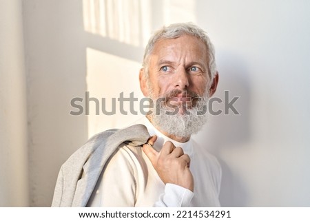 Thoughtful mature older gray-haired business man leader, pensive middle aged senior professional businessman wearing suit looking through window thinking standing at white wal in sunlight. Royalty-Free Stock Photo #2214534291