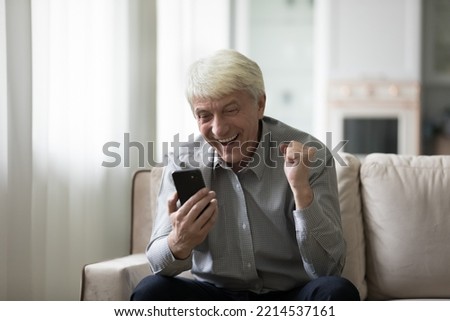 Overjoyed older man sits on sofa holding smartphone celebrate amazing news, relish success, received great commercial offer, betting win, moment of on-line gambling, auction or lottery victory, luck Royalty-Free Stock Photo #2214537161