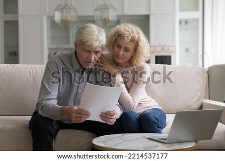 Concerned older couple, wife and husband sit on sofa at home read received formal notification from bank about mortgage denial, unpaid taxes, financial issues, learn insurance terms looking serious Royalty-Free Stock Photo #2214537177