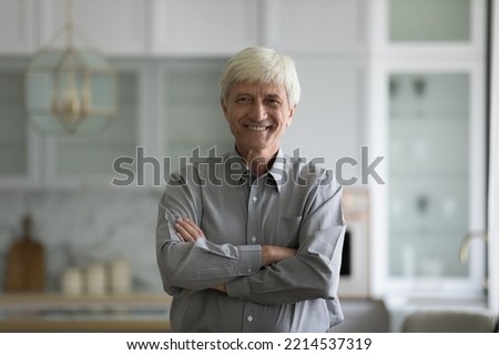 Retired grey-haired man standing in fashionable domestic kitchen with arms-crossed smile look pose for camera. Happy homeowner, wellbeing carefree life on retirement, optimistic older person portrait Royalty-Free Stock Photo #2214537319