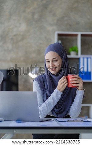 A beautiful Asian Muslim woman wears a headscarf using her laptop to do financial accounting work in her living room at home.