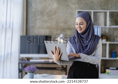 A beautiful Asian Muslim woman wears a headscarf using her laptop to do financial accounting work in her living room at home.