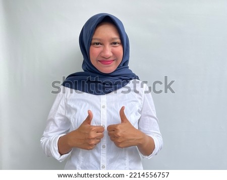 Portrait of smiling young asian woman making okay or good signs with thumbs up. Indonesian woman wearing white shirt and navy hijab isolated by white background