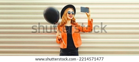 Portrait of happy smiling young woman taking selfie with smartphone and black balloon wearing round hat, red jacket on background