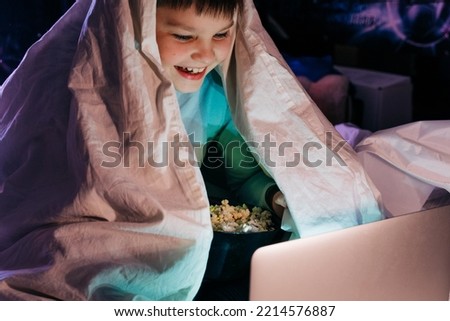 Happy kid son laughing eat popcorn remote control watching funny comedy tv show sitting on sofa having fun viewing video on laptop in evening at home. Teenager gen z boy under blanket at Movie night