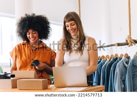 Cheerful businesswomen using a laptop while preparing an online order in a thrift store. Happy small business owners making plans for a shipment. Female entrepreneurs running an online clothing store. Royalty-Free Stock Photo #2214579713
