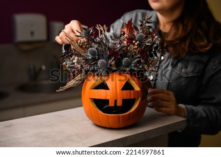 hand of woman florist gently decorate lovely autumn arrangement of thistles flowers and plants in bright orange pumpkin with carved eyes and mouth. Beautiful floral decor for halloween. Close-up
