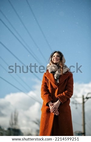 Young brunette girl in orange coat with scarf around her neck, smiling, stands with her palms crossed under falling snow, looking away, against background of blurred power lines, with copy space.