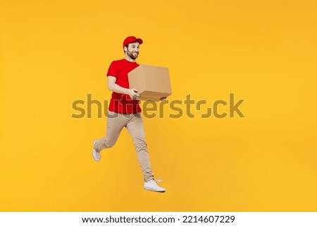 Full body side view fun delivery guy employee man in red cap T-shirt uniform workwear work as dealer courier jump high hold cardboard box run fast isolated on plain yellow background. Service concept Royalty-Free Stock Photo #2214607229