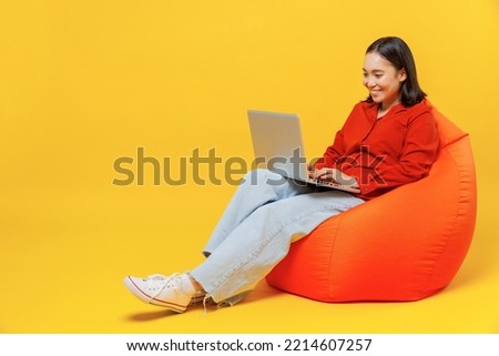Full size body length smart young woman of Asian ethnicity 20s years old wears casual clothes sit in bag chair hold use work on laptop pc computer isolated on plain yellow background studio portrait