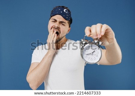 Young sleepy tired sad yawning caucasian man 20s wear pajama jam sleep mask resting relax at home holding in hand clock alarm isolated on dark blue background studio. Bad mood night bedtime concept.