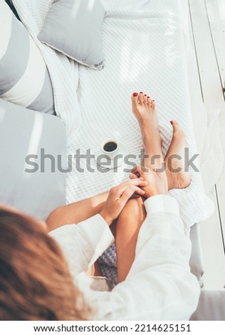 Beautiful barefoot woman dressed white shirt relaxing on luxury terrace with espresso cup patio on comfortable sofa pillows, enjoying midday time. Carefree life or coffee break concept top view image.