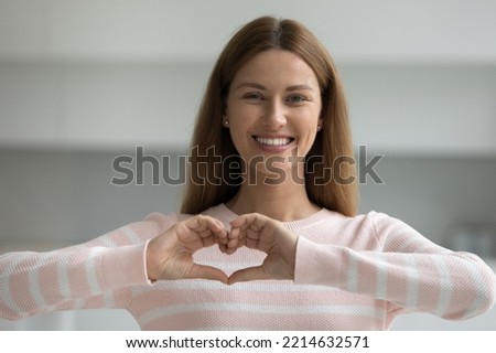Beautiful woman showing heart sign with folded fingers smiling looking at camera, head shot portrait. Attractive optimistic young female demonstrate symbol of love feels happy posing alone at home