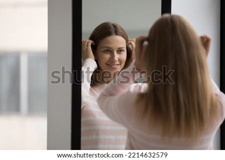 Young beautiful woman in casual sweater, preening, do morning routine, touch her hair standing in front of mirror smiling admire herself feels satisfied with appearance and complexion. Beauty, fashion Royalty-Free Stock Photo #2214632579