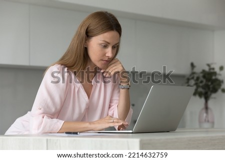 Attractive serious woman working on laptop, learns new software, doing telework busy in online task seated in kitchen. Female making order distantly, buying on internet, choose services looks pensive