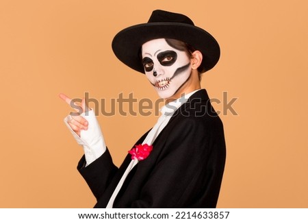 Smiling man dresses for carnival party indicates away on blank space wears halloween costume and bright makeup isolated on beige background. Street performer