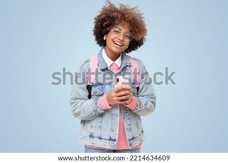 Funny african american laughing school girl with afro hairstyle and trendy glasses holding phone, looking at camera, isolated on blue background