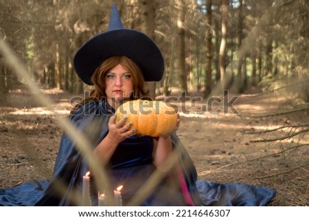 A young red-haired witch in a cloak and a pointed hat with a pumpkin in her hands in the forest. horizontal photo