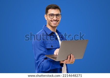 Portrait of young modern business man standing in casual blue shirt, holding laptop and looking at camera with happy smile, isolated on blue