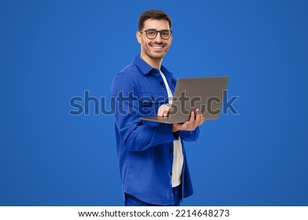 Portrait of young modern businessman standing holding laptop and looking at camera with happy smile, isolated on blue