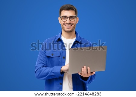 Studio portrait of young man standing holding laptop and looking at camera with happy smile, isolated on blue