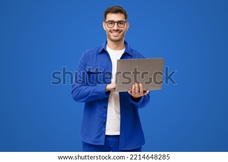 Young man standing in casual blue shirt, holding laptop and looking at camera with happy smile, isolated on blue background