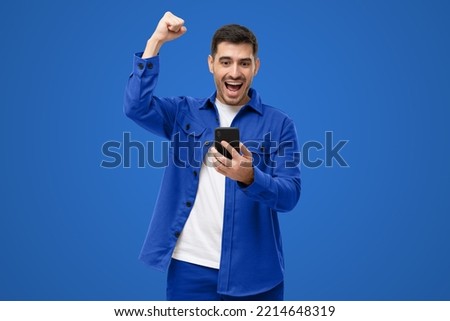 Happy excited sucessful modern man holding phone and raising arm up to celebrate achievement, isolated on blue background