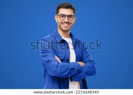 Young hispanic man wearing blue shirt and glasses, looking at camera with positive confident smile, holding arms crossed, isolated on blue background Royalty-Free Stock Photo #2214648345