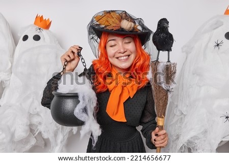 31st of October. Satisfied young woman with orange hair smiles gently keeps eyes closed holds caulderon broom prepares for holiday celebration wears hat and black dress poses near spooky ghosts