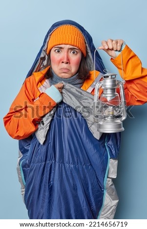 Vertical shot of displeased young Asian woman has red frozen face holds lantern to see in darkness frowns face looks unhappily poses in sleeping bag spends night in nature has adventure trip