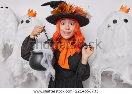 Happy holiday concept. Lovely Asian woman licks lips holds caulderon wears witch hat black dress and orange bow around neck poses against scary decor prepapares for Halloween frightens during night