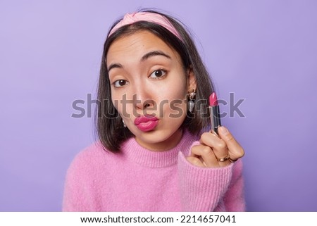 Portrait of pretty Asian woman pouts lips holds lipstick applies makeup wears headband and pink jumper enjoys her beauty and face care routine isolated over purple background promots cosmetic product