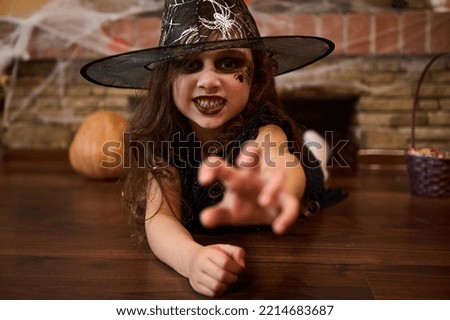 Scary child girl, a sorceress in a wizard's hat, stretches out her hand, looks at the camera with a frightening look, lies on the floor near a brick wall covered with cobwebs, and Halloween decoration