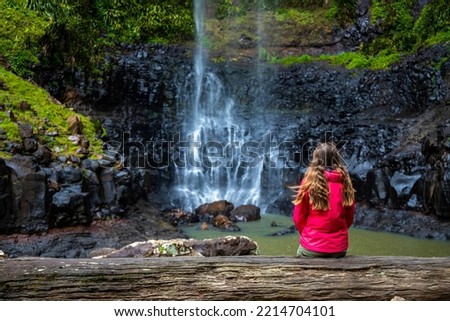 a long-haired girl stands beneath a powerful tropical waterfall in springrbook national park, australia; hiking through the jungle in queensland