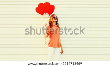 Portrait of happy smiling young woman with bunch of red heart shaped balloons wearing shirt, sunglasses on white background