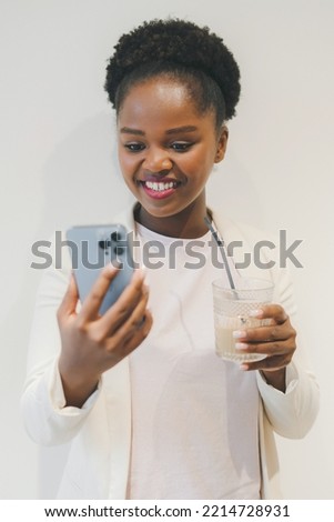 Young pretty african woman typing message on smart phone and holding a coffee cup hand while standing isolated over white cafe wall. African girl looking at the Royalty-Free Stock Photo #2214728931