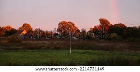 Village evening landscape with autumn trees and green field under rainbow