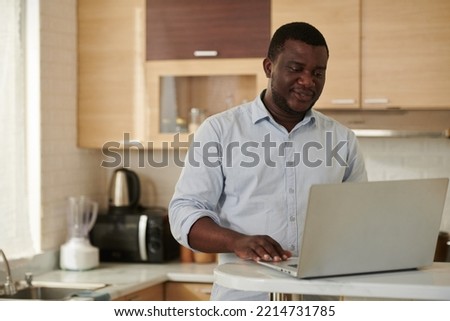 Smiling businessman standing at kitchen counter when having online meeting with colleagues